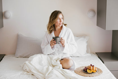 Femme portant un peignoir blanc dans un lit. Elle a dans ses mains une tasse à café. Sur le lit blanc il y a un plateau de fruit